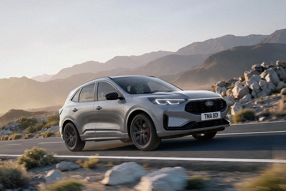 Une Ford Kuga gris argenté roulant sur une route de montagne entre des rochers et des chaînes de montagnes.