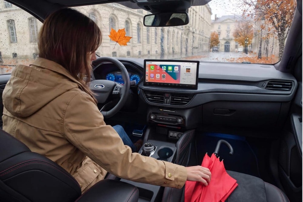 Une photo de l'intérieur d'un véhicule Ford en automne. Une femme vêtue d'une veste marronne est assise au volant et tend la main vers un parapluie rouge posé sur le siège passager.