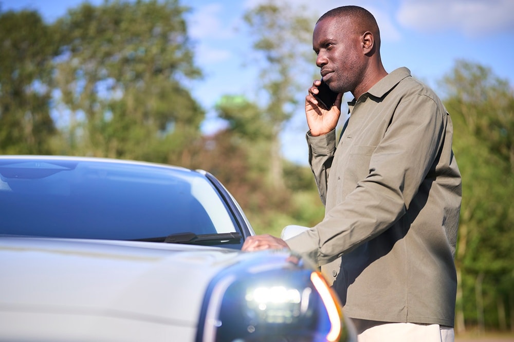 Une personne téléphone à côté d'une voiture de couleur claire en extérieur.