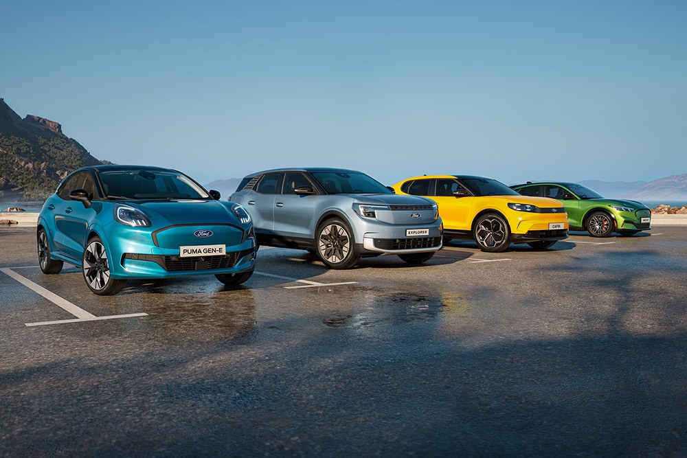 Quatre voitures électriques Ford de couleurs différentes garées côte à côte, avec des montagnes et un ciel bleu clair en arrière-plan.