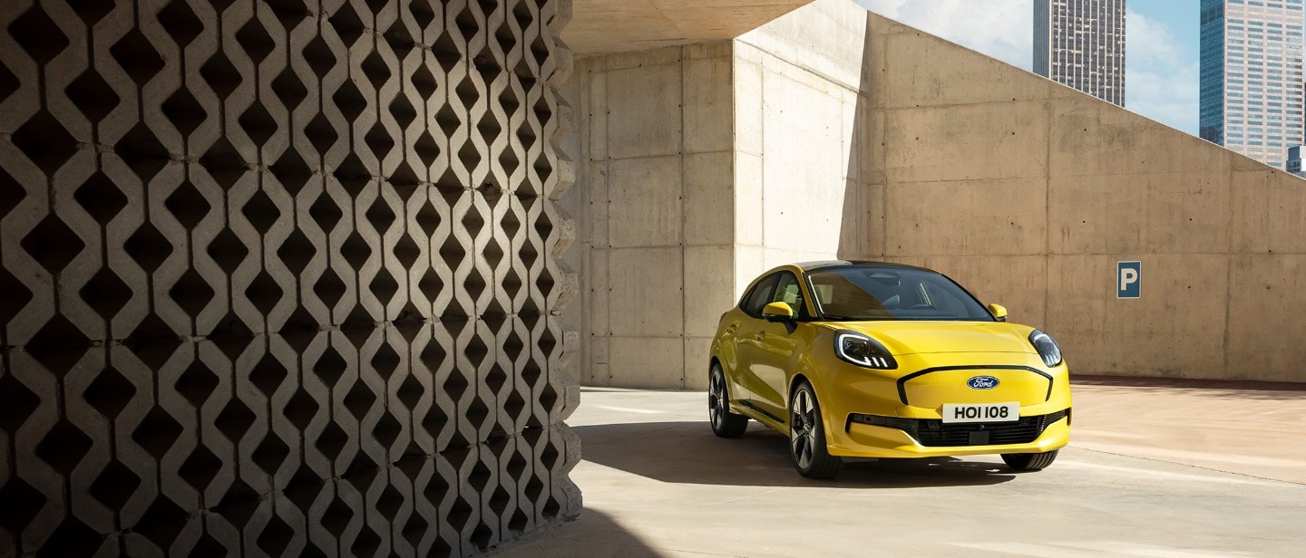 Une Ford Puma Gen-E jaune vif est garé sur une surface en béton sous une architecture moderne et angulaire en béton.