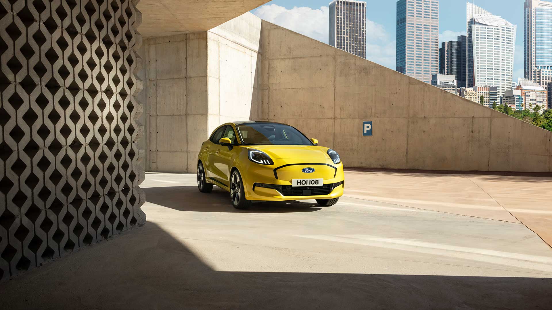 Une Ford Puma Gen-E jaune vif est garé sur une surface en béton sous une architecture moderne et angulaire en béton.