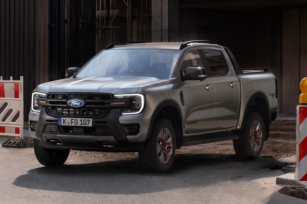 Ford Ranger gris argenté à quatre portes avec benne ouverte, garé devant un bâtiment à façade vitrée sombre.