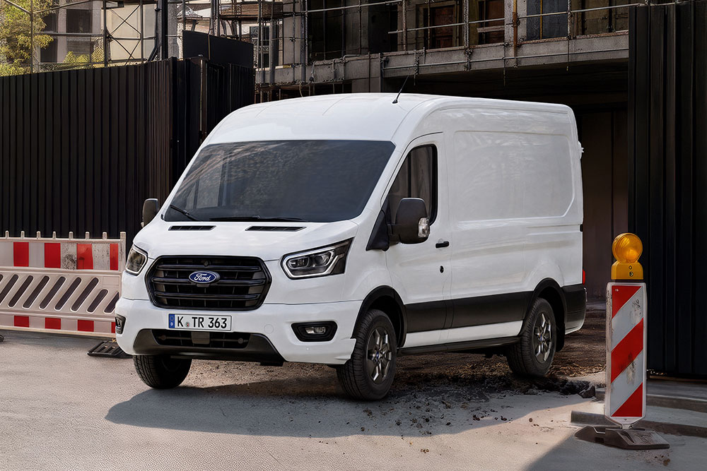 Ford Transit blanc à toit surélevé et habillage de cabine sombre, garé devant une façade sombre avec une barrière rayée rouge et blanc.