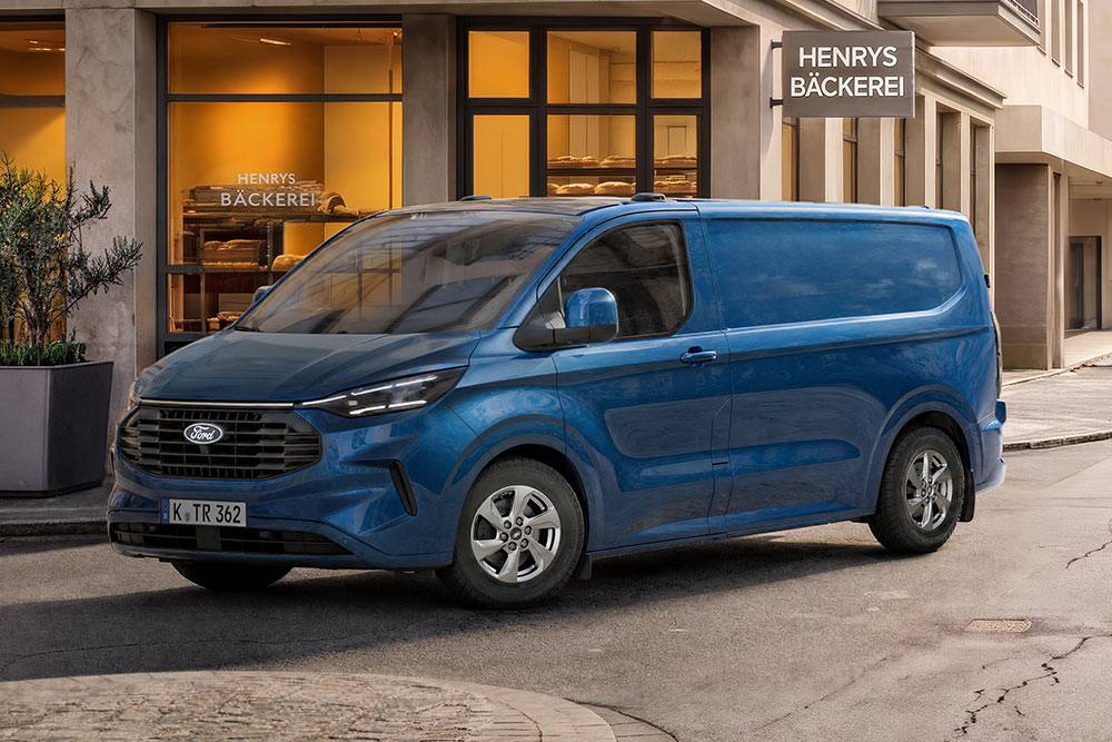 Ford Transit Custom bleu vif garé devant un bâtiment de couleur claire aux grandes fenêtres, avec en arrière-plan une enseigne indiquant 'Henrys Bakery'.