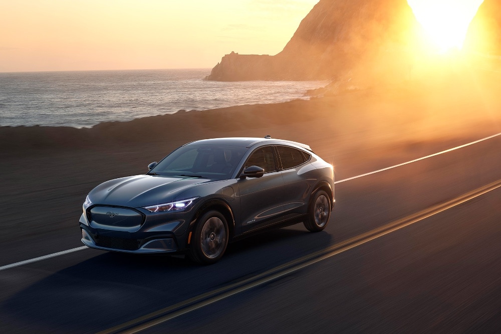 Une voiture électrique Ford Mustang Mach-E roule sur une route côtière au coucher du soleil.