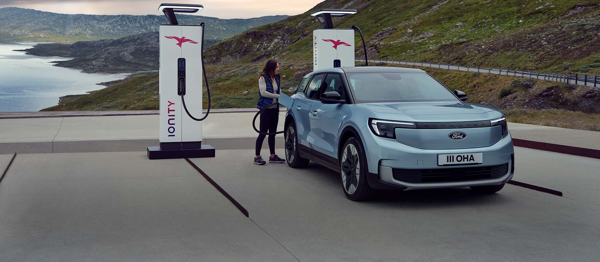 Une femme branche un Ford Explorer bleu, 100% électrique, à une borne de recharge publique à l'aide d'un câble. La voiture est garée près de montagnes et d'eau.