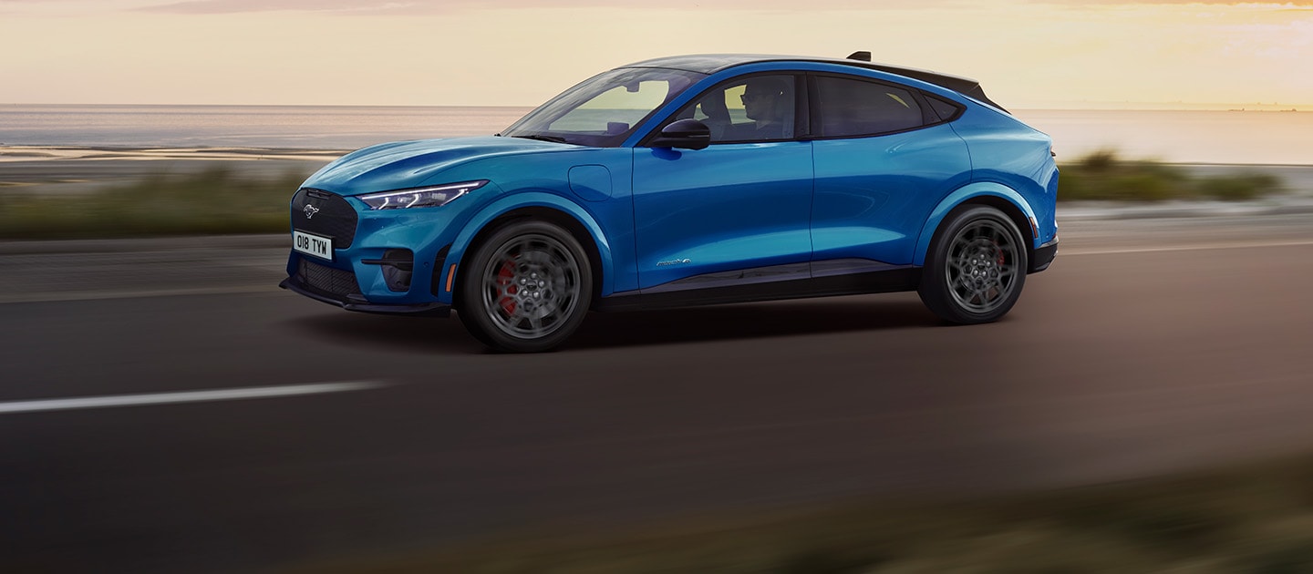 Un Ford Mustang Mach-E électrique bleu roulant le long d'une route côtière au coucher du soleil.