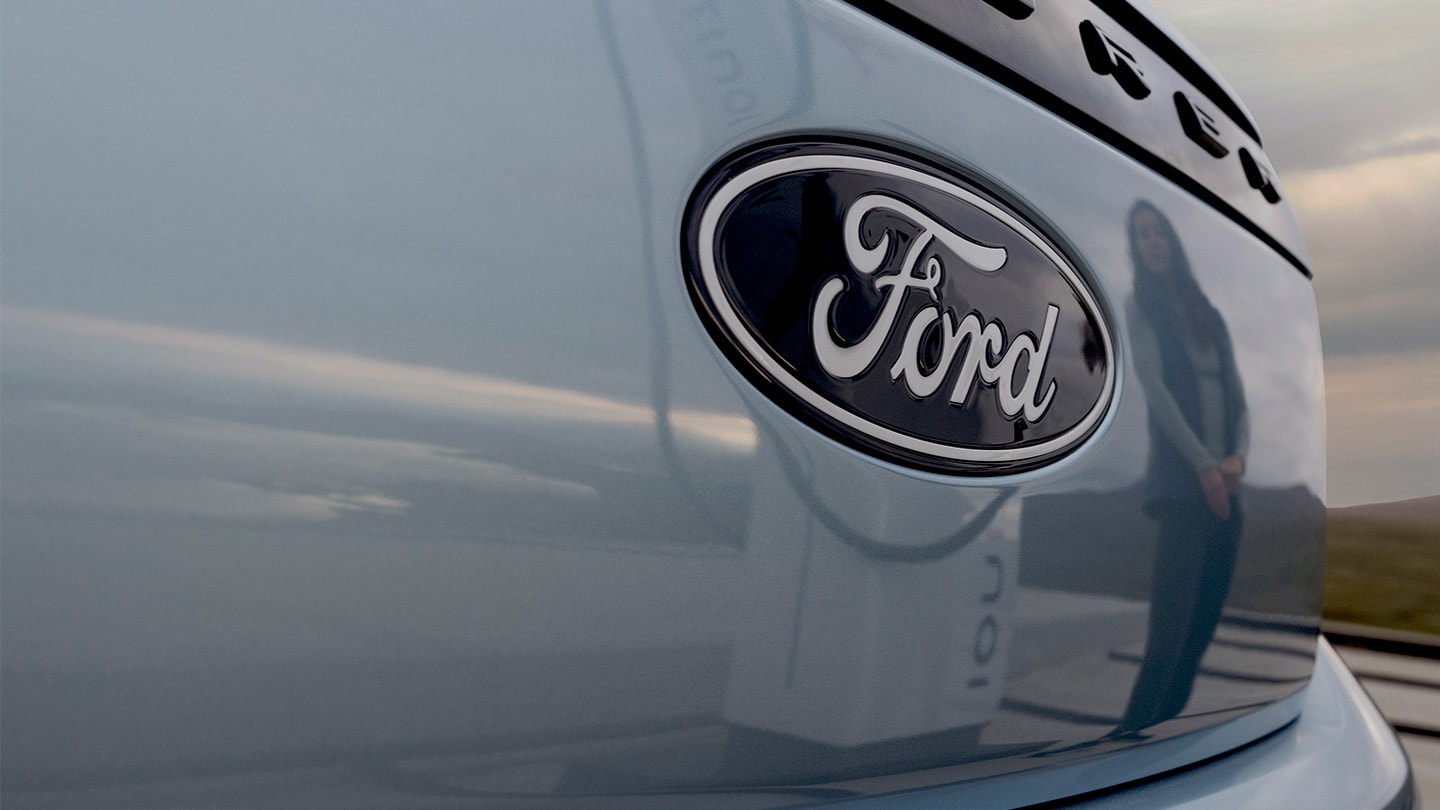 Logo Ford sur un Ford Explorer électrique bleu avec une station de recharge publique reflétée dans la voiture.
