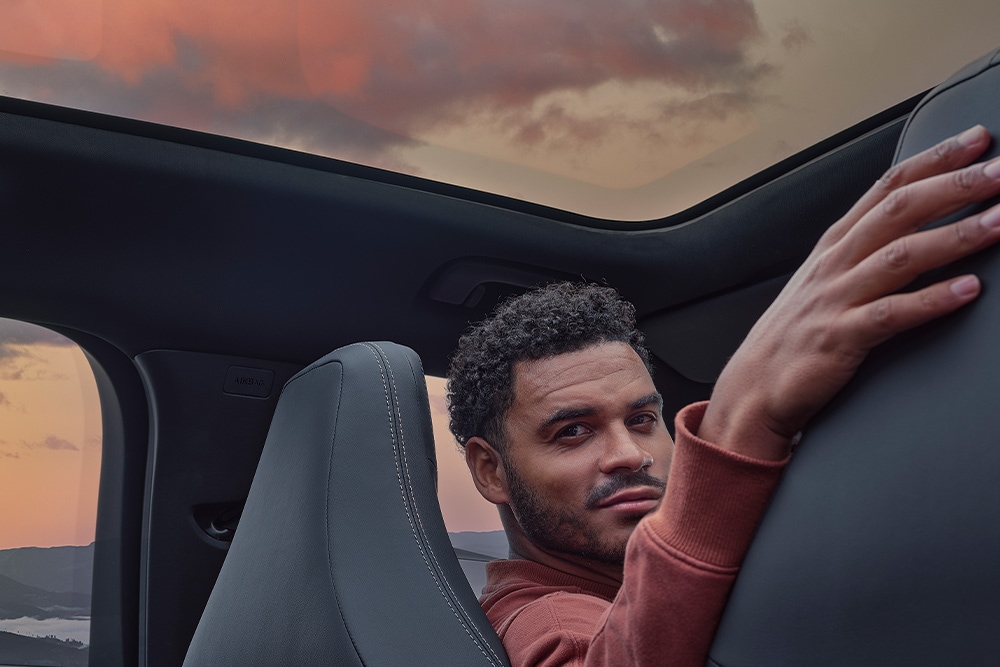 Un homme assis dans une voiture au coucher du soleil, la main posée sur le dossier d'un siège.