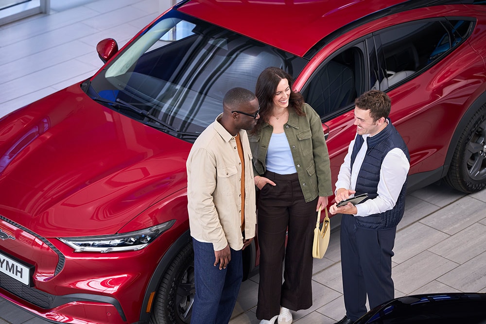 Un couple souriant discute avec un vendeur Ford dans un showroom, à côté d'un Ford Mustang Mach-E rouge.
