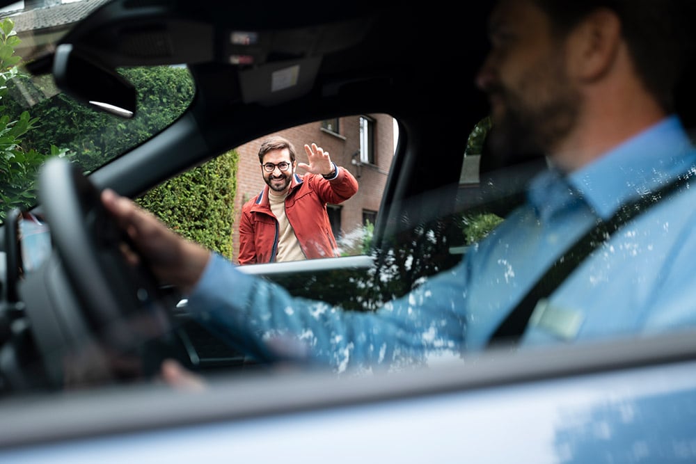 Un homme au volant d'une voiture salue un autre homme souriant à l'extérieur.