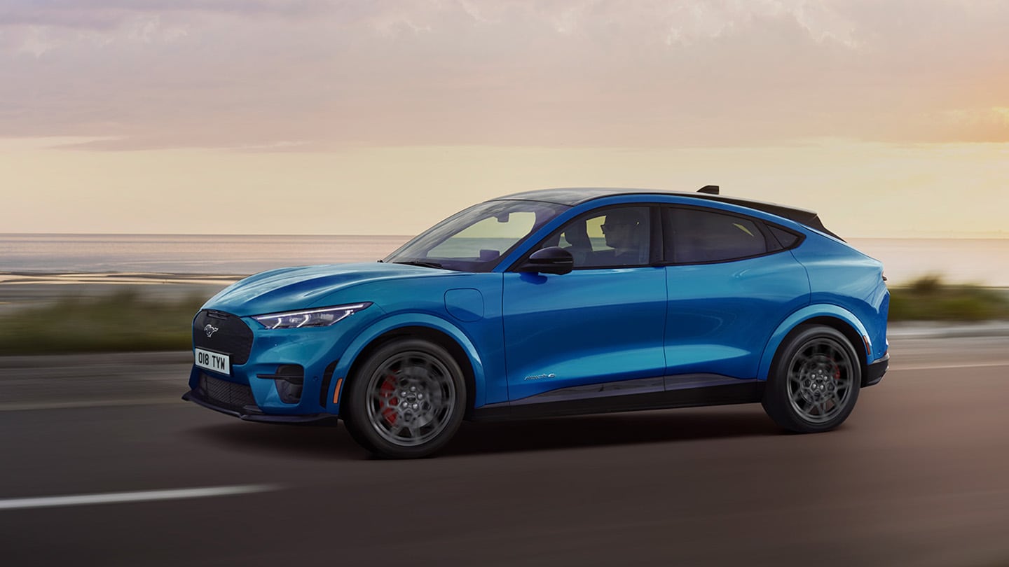 Vue latérale d’une Ford Mustang Mach-E électrique bleue roulant le long d’une route côtière au crépuscule.