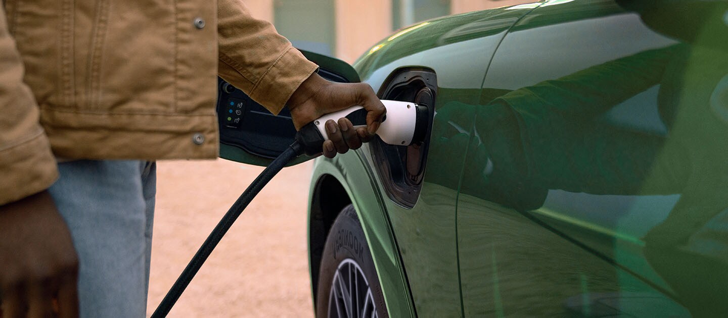 Une personne vêtue d’une veste crème branchant un câble de recharge électrique sur sa Ford Mustang Mach-E électrique verte.