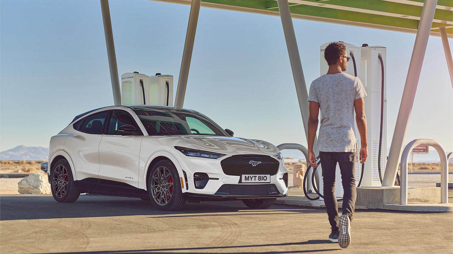 Ford Mustang Mach-E blanche garée à une station de recharge publique en extérieur. Un homme en t-shirt blanc se dirige vers la voiture.