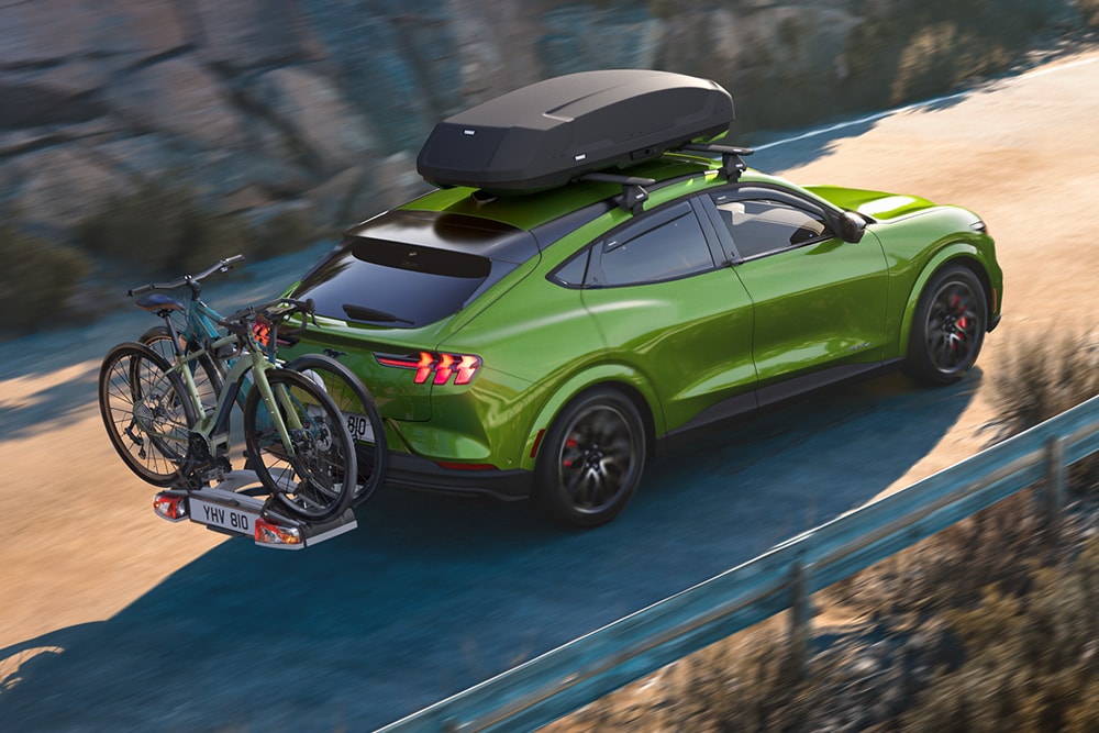 Vue arrière d'une Ford Mustang Mach-E verte, équipée d'un coffre de toit et d'un porte-vélos, circulant sur une route pittoresque.