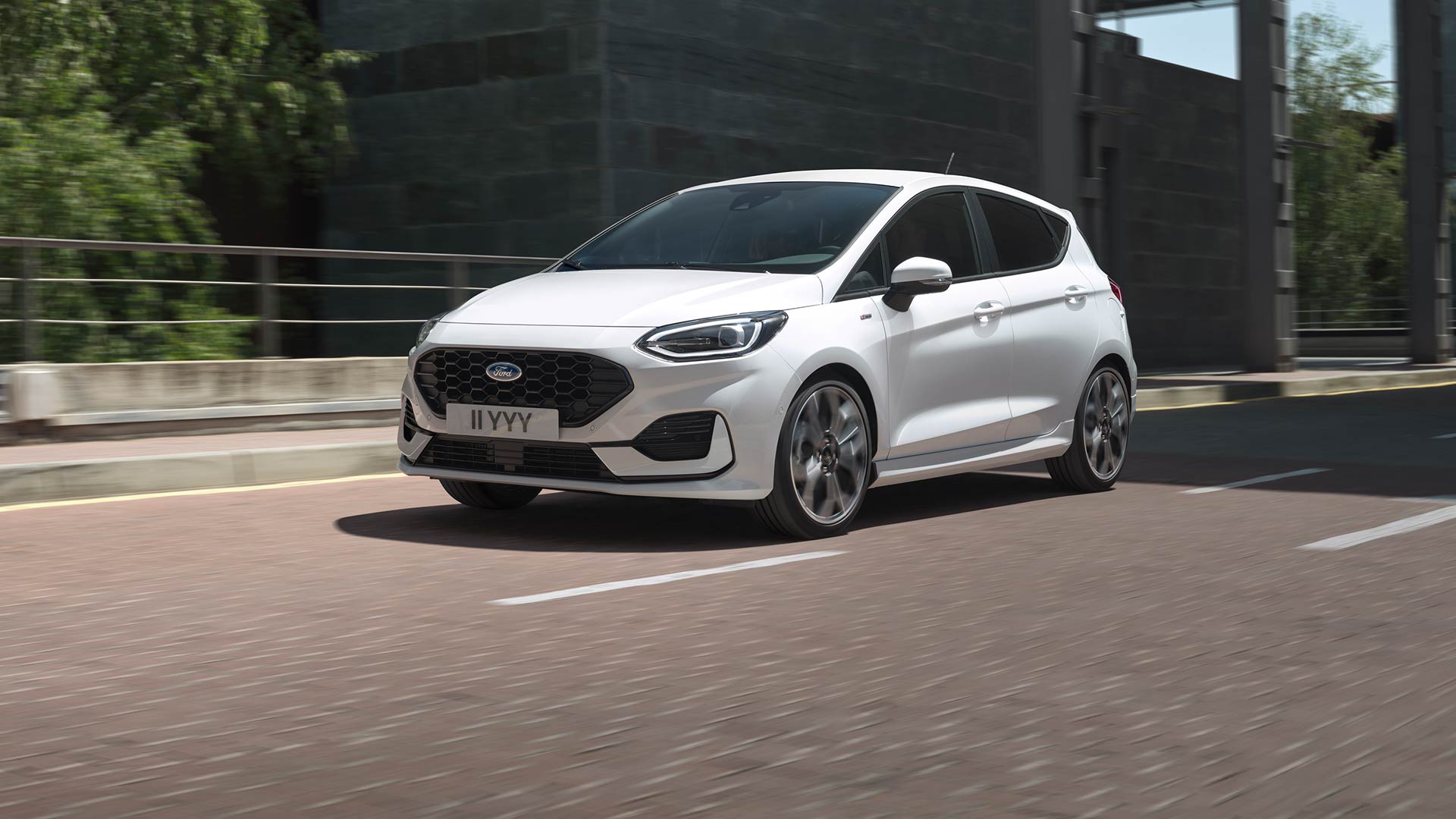 Ford Fiesta blanche roulant sur une route urbaine, avec de la verdure et des bâtiments en arrière‑plan.