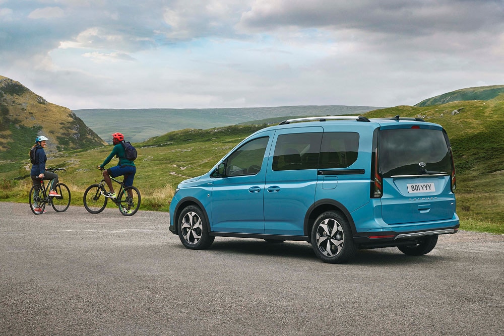 Une Ford Tourneo Connect bleue est garée sur une route dans un paysage verdoyant et vallonné, à proximité de deux cyclistes.