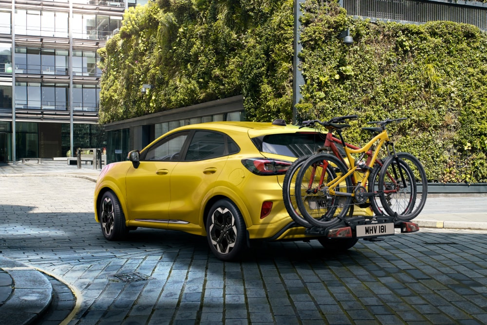 Ford Puma Gen-E jaune garé dans une zone urbaine avec deux vélos sur un porte-vélos à l'arrière.