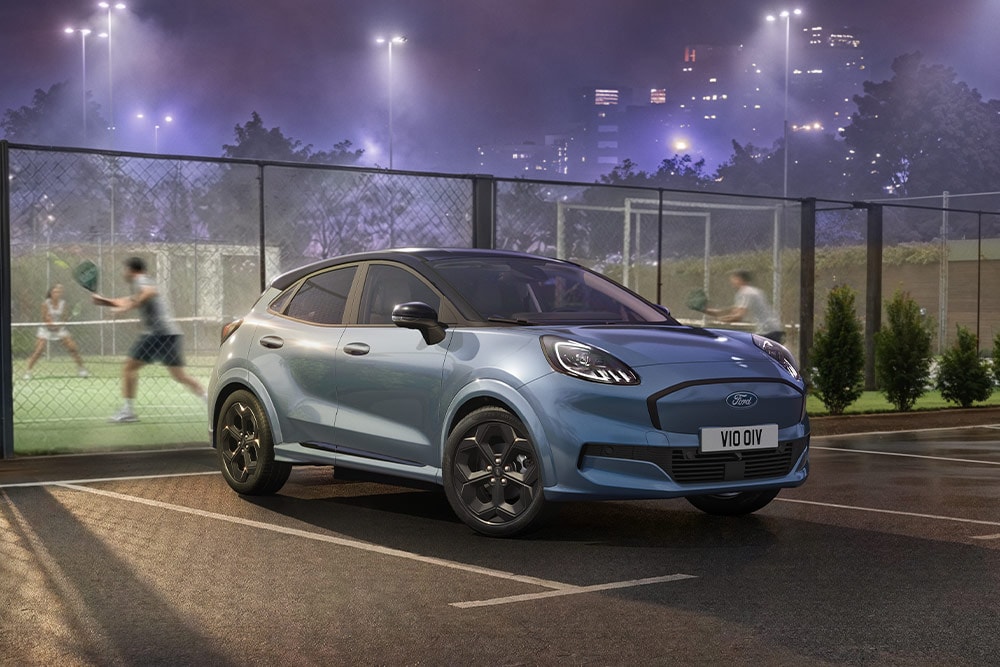 Un Ford Puma Gen-E bleu garé à côté d’un court de tennis, avec les lumières de la ville en arrière-plan.