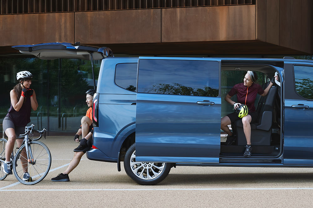 Ford Tourneo Custom bleu stationné avec les portes latérales ouvertes et des personnes avec des vélos à proximité. 