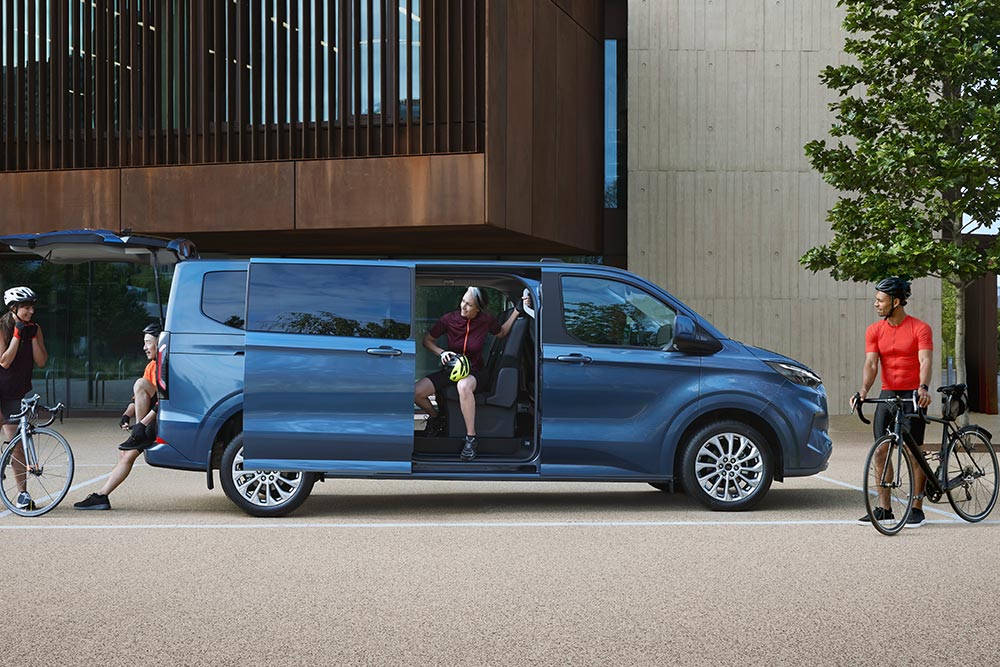 Fourgon Ford Tourneo Custom bleu stationné avec les portes latérales ouvertes et des personnes avec des vélos à proximité. 
