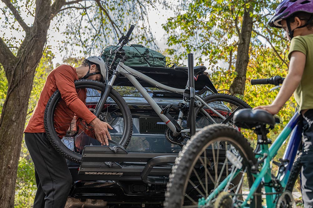 Une personne charge un vélo sur le porte-vélos arrière d'une voiture, avec un enfant et un autre vélo à proximité.