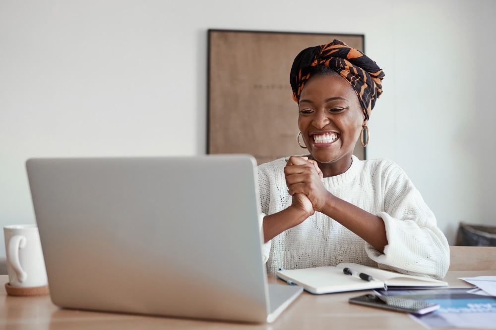 Une personne à l'expression joyeuse regarde un ordinateur portable, les mains jointes d'excitation à un bureau.