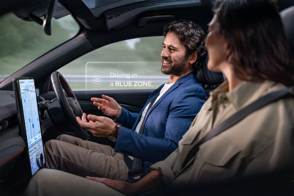 Un homme et une femme dans une Mustang Mach-E conduisent sans les mains avec Ford BlueCruise. Une notification indique "Conduite dans une BLUE ZONE ".