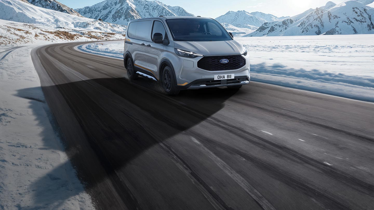 Un Ford E-Transit Custom argenté roulant sur une route de montagne enneigée sous un ciel bleu clair.