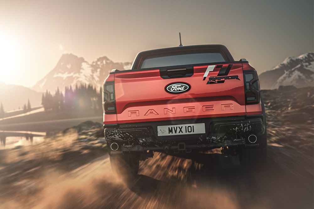Vue arrière d'un Ford Ranger Raptor rouge roulant sur un chemin de terre, avec des montagnes en arrière-plan.