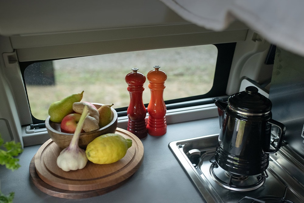Bloc cuisine intégré du Ford Transit Custom Nugget, avec une bouilloire et des fruits posés sur le plan de travail.