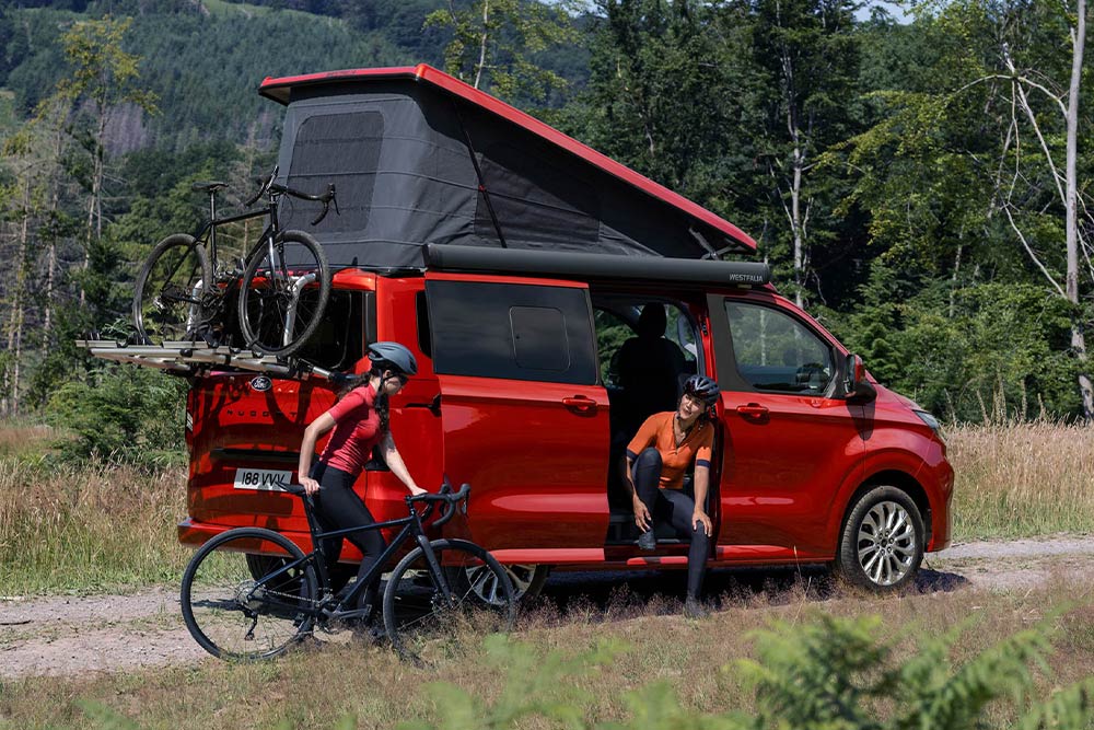 Des personnes déchargent des vélos du porte-vélos d’un Ford Transit Custom Nugget rouge.