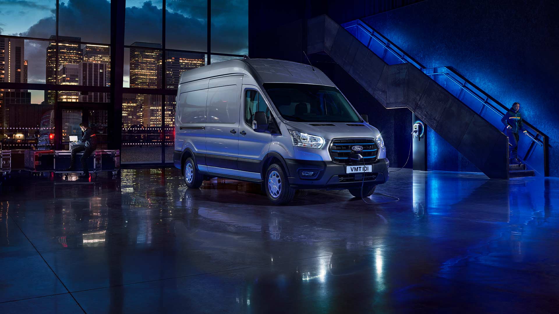 Fourgon électrique Ford E‑Transit argenté garé à l’intérieur, avec une vue sur la ligne d’horizon de la ville à travers de grandes fenêtres. 