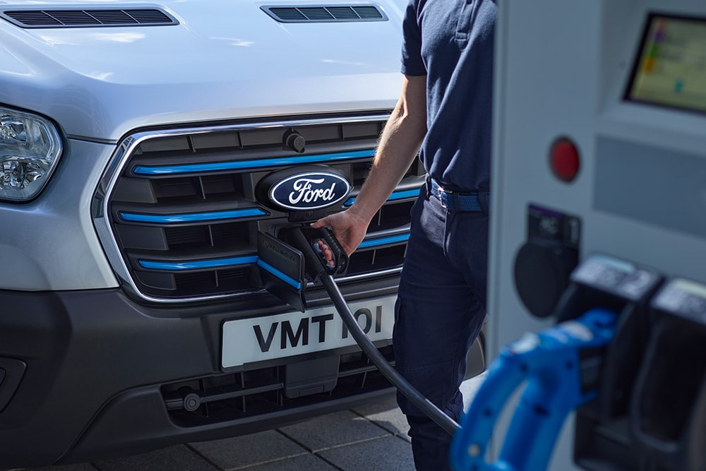 Gros plan d’une personne branchant un câble de recharge sur un fourgon électrique Ford E‑Transit argenté.