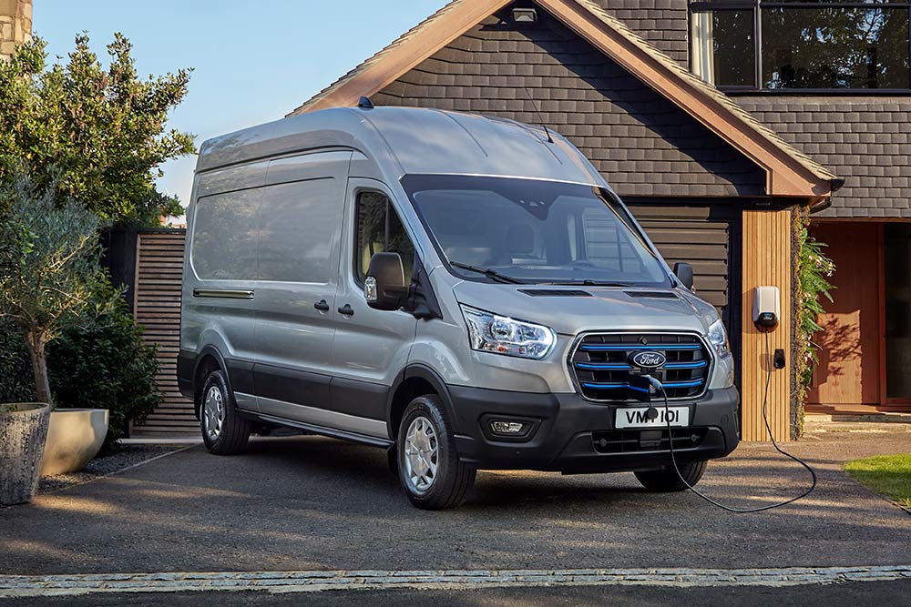 Fourgon Ford E‑Transit argenté garé dans une allée à côté d’une maison, branché à une borne de recharge.