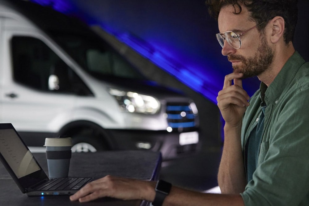 Un homme travaillant sur un ordinateur portable avec un fourgon électrique Ford E‑Transit à l’arrière-plan.