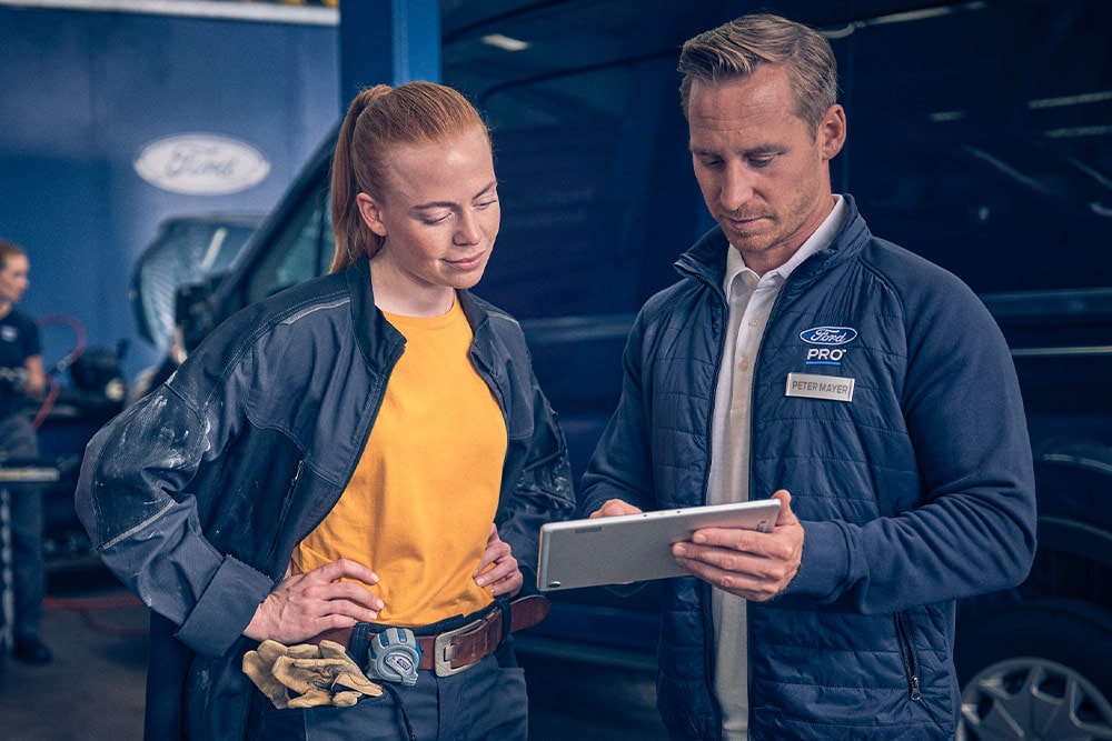 Deux mécaniciens dans un garage regardant une tablette devant un fourgon électrique Ford E‑Transit.