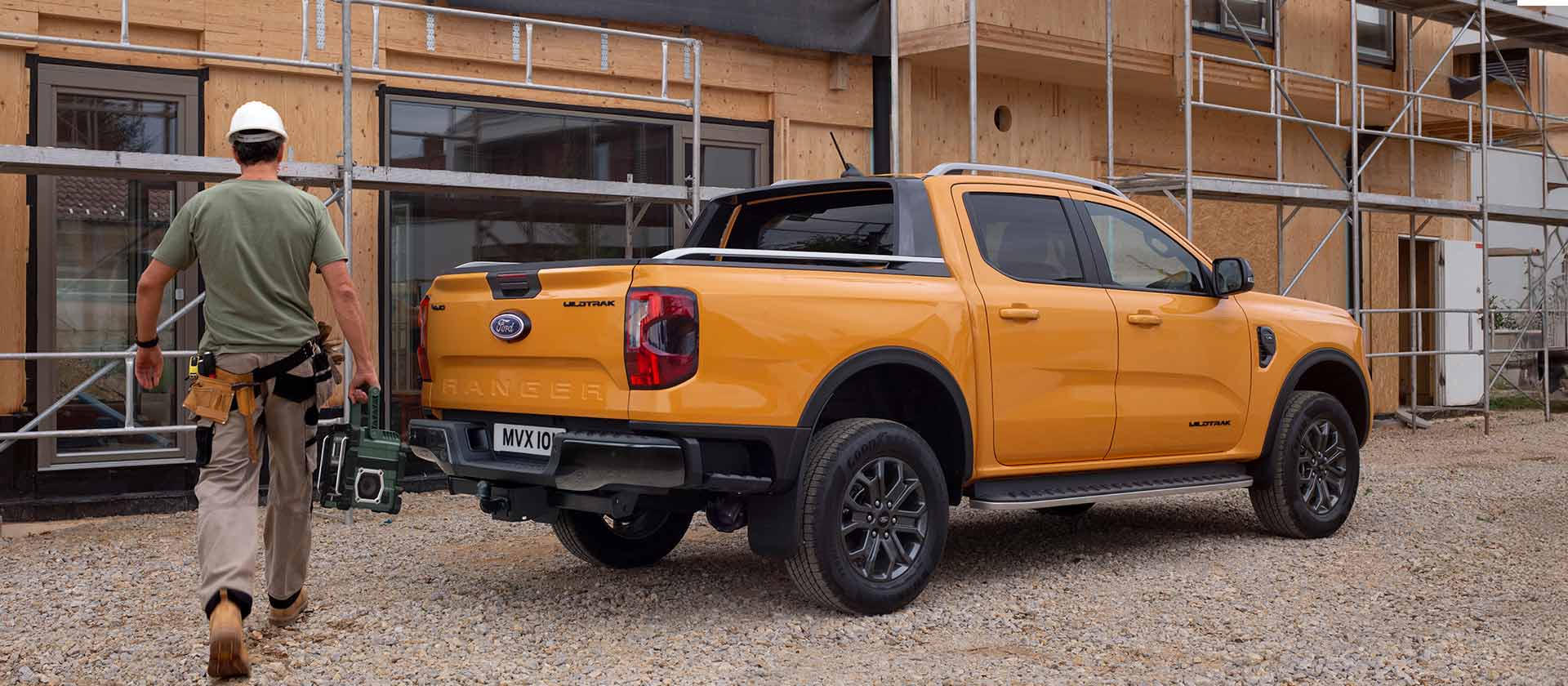Un travailleur portant un casque de sécurité marche vers un pick-up Ford Ranger jaune sur un chantier de construction.