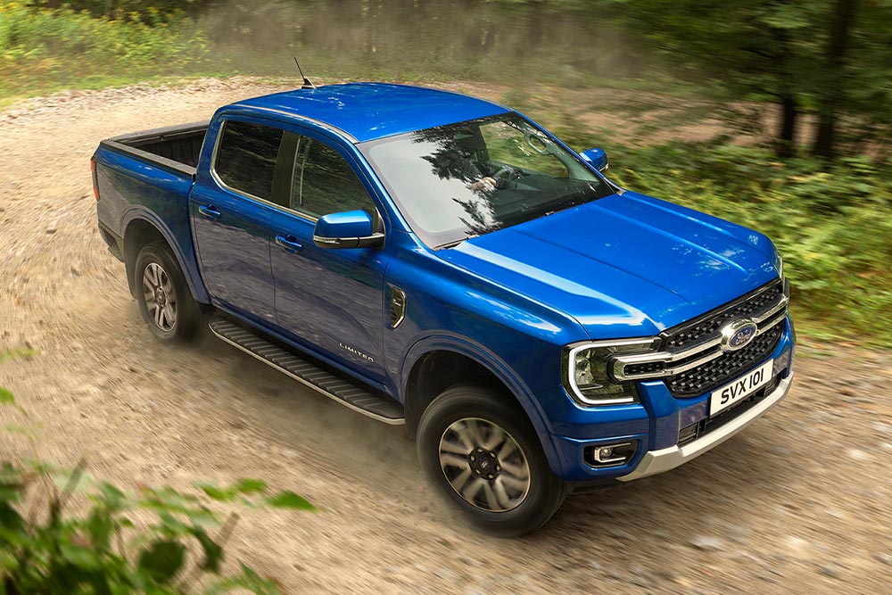 Un pick-up Ford Ranger bleu roule le long d'un chemin de terre à travers une forêt, laissant une traînée de poussière.