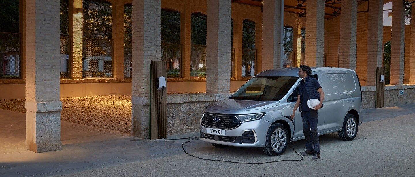 Un homme qui tient un casque de sécurité branche un câble de chargement d’un boîtier mural dans le nouveau Ford Transit Connect en Stardust Silver.