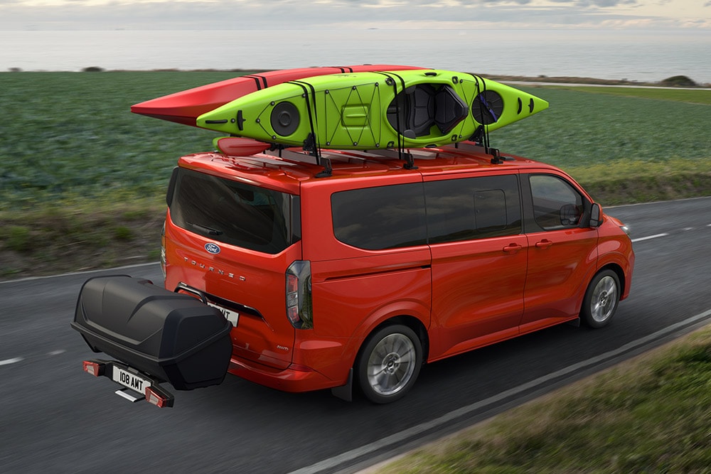 Ford Pro Transit rouge avec deux kayaks sur le toit, remorquant un porte-bagages, et roulant le long d'une route côtière.