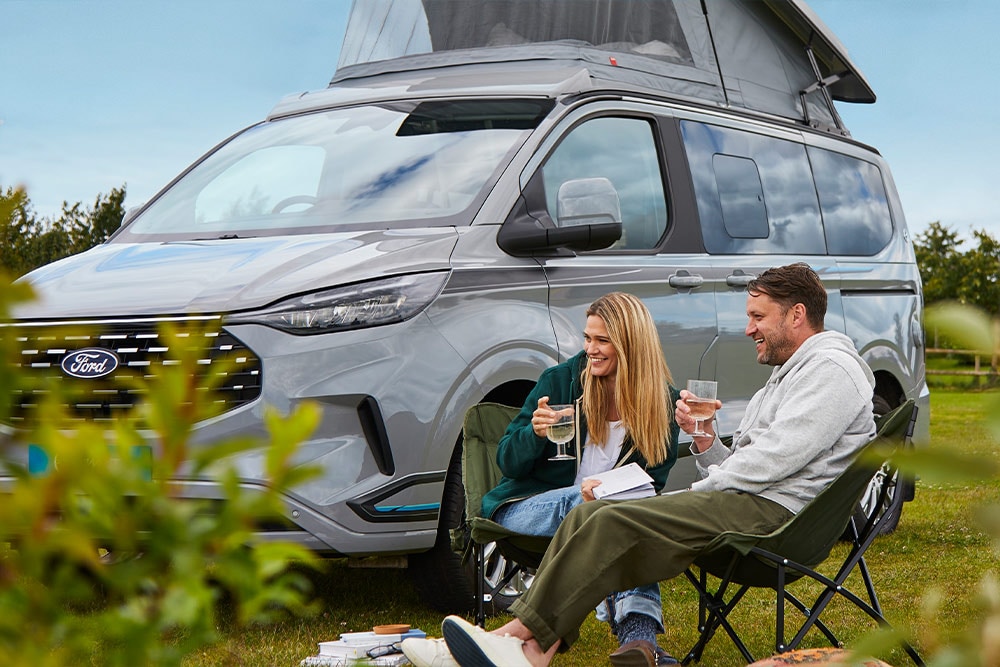 Un couple savourant des boissons dans des chaises de camping à côté d'un camping-car Ford Transit Custom gris avec un toit relevable.