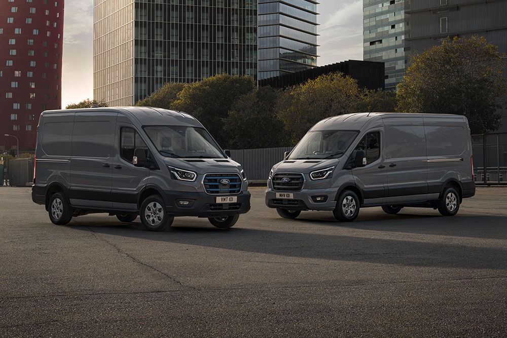 Deux fourgons Ford Pro Transit, dont un électrique, garés sur un parking avec des bâtiments modernes en arrière-plan.