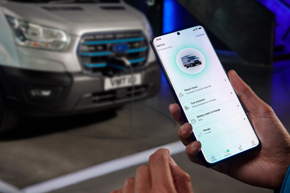 Une personne tient un smartphone affichant une application Ford, avec un Ford E-Transit Custom en charge à l'arrière-plan.