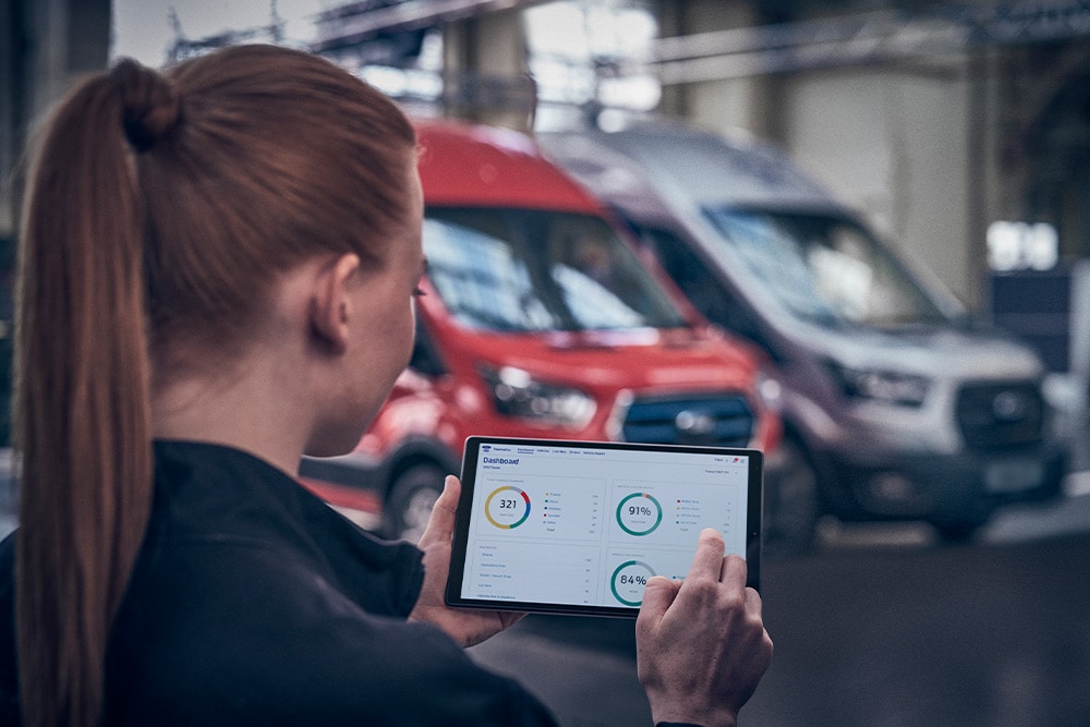 Une femme regarde une tablette affichant des tableaux de bord, devant des camionnettes garées.