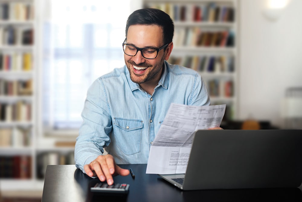 Un homme à lunettes sourit en utilisant une calculatrice et en tenant des documents à un bureau avec un ordinateur portable.
