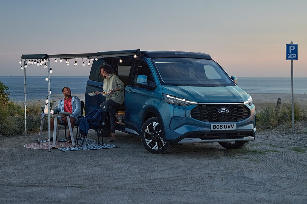 Deux campeurs se détendent près d'un Ford Pro Transit Custom Nugget bleu, une zone de stationnement en bord de mer en arrière-plan.