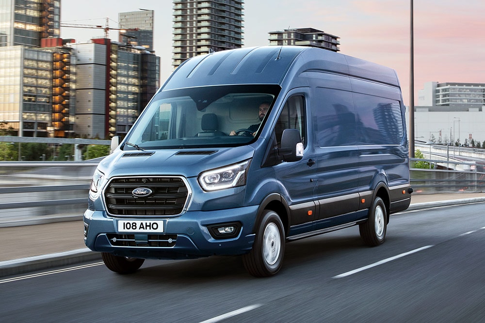Un Ford Transit argenté circule dans un paysage urbain.