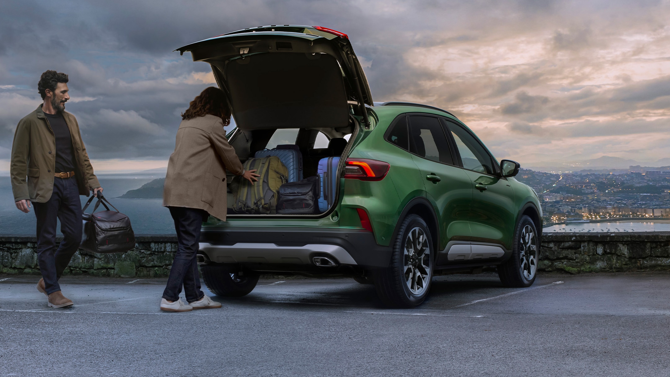 Un homme et une femme chargent des bagages dans le coffre d'un SUV vert garé sur un belvédère pavé.